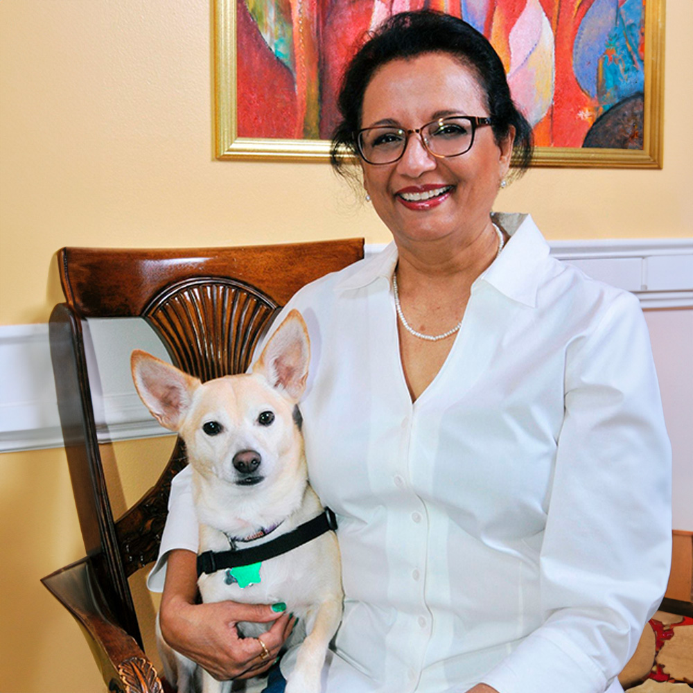 A woman wearing glasses, a white shirt, and a black vest is sitting on a chair holding a small dog.
